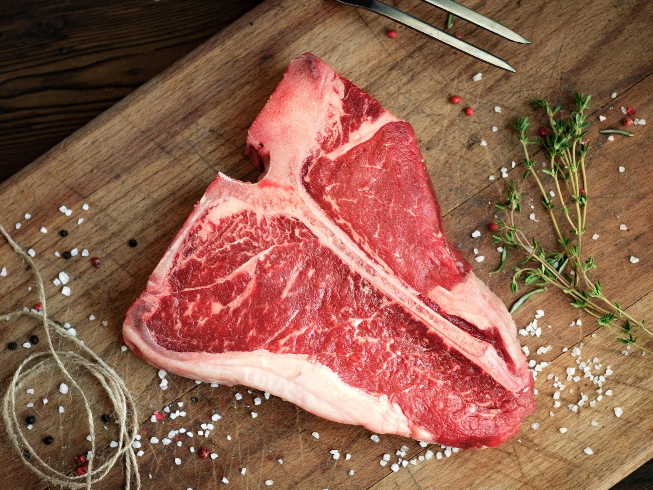 beef_tbone_raw Fresh T-bone steak on a wooden cutting board, surrounded by coarse sea salt, black and pink peppercorns, and fresh thyme, ideal for grilling or cooking.