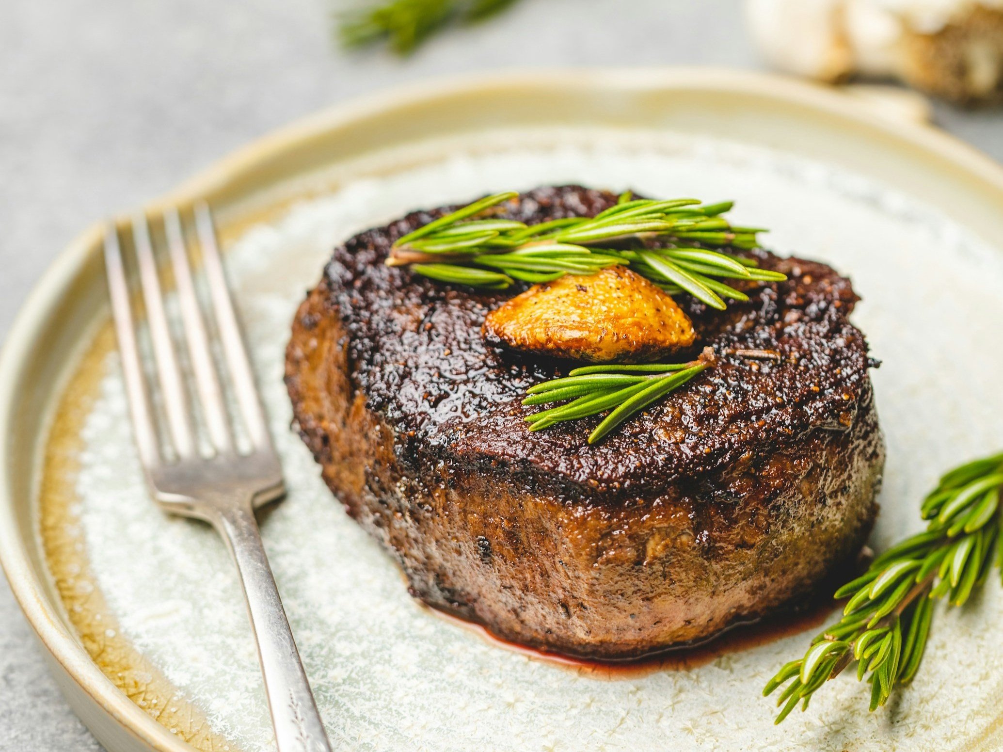 Juicy grilled filet mignon topped with herb butter and fresh rosemary, served on a textured plate with a fork, showcasing a gourmet dining experience.